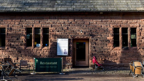Exterior view of the brick restaurant building, featuring a sign announcing "Restaurant. Delicious Home Made Food," a historical plaque, dark windows, a doorway and a child's stroller. Outdoor seating is situated nearby.